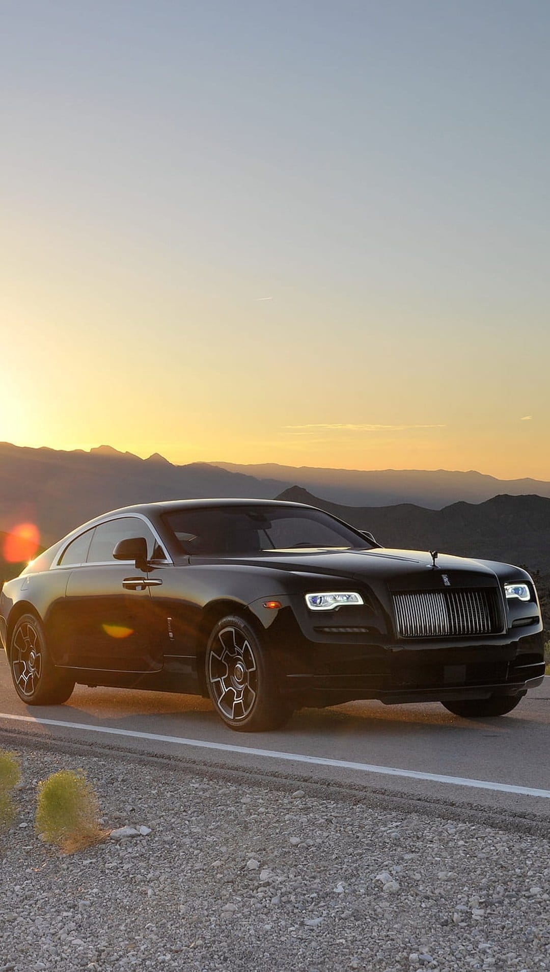 Black Rolls-Royce coupe driving on a desert road against orange sunset mountains and a golden sky background.