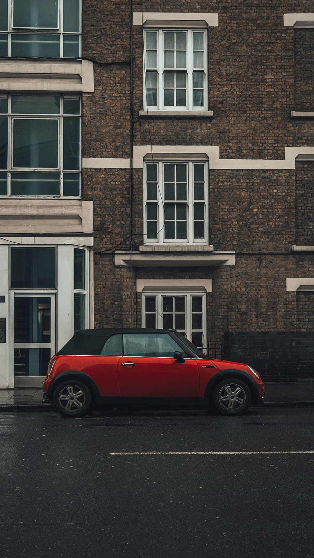 Red Mini Cooper convertible parked on wet asphalt next to a brick building with traditional white sash windows.