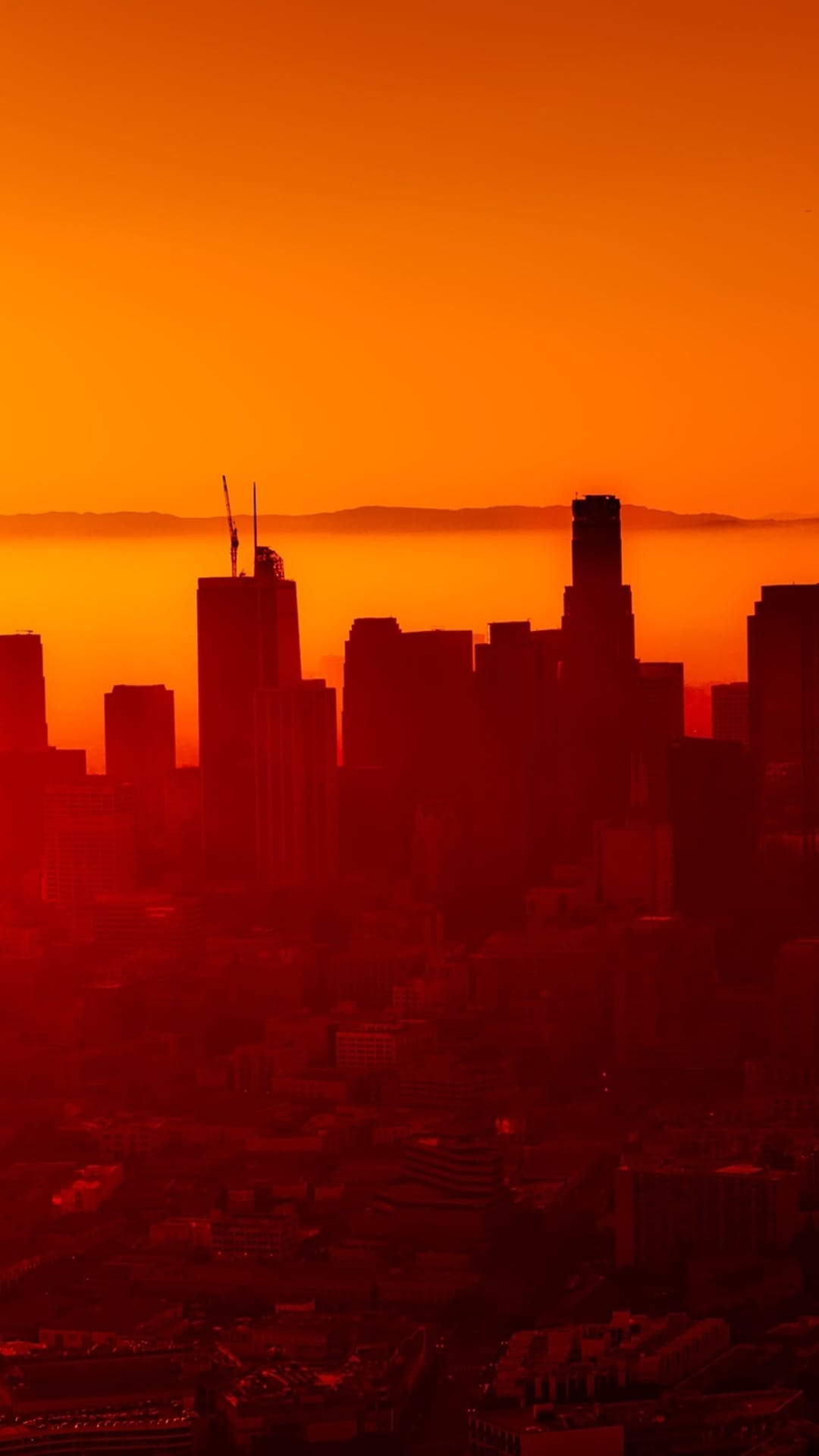 Black city buildings silhouetted against a glowing red and orange sky with hazy mountain peaks in the distance.