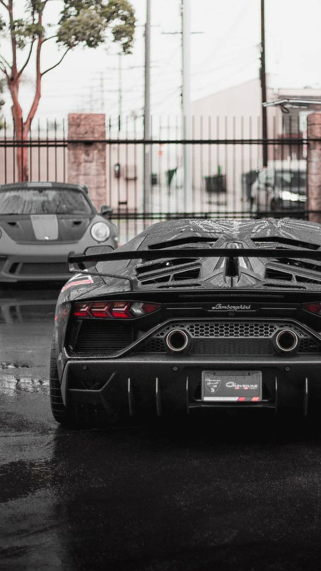 Black Lamborghini Aventador with glowing red taillights parked behind a grey Porsche 911 on a wet asphalt street.