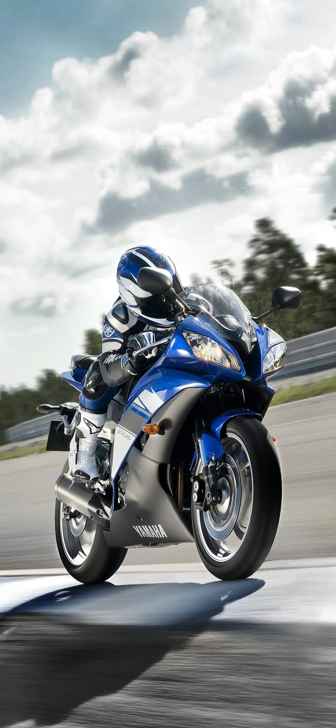 Yamaha motorcycle racer leans into a track corner wearing full safety gear on a blue sportbike under clouds.