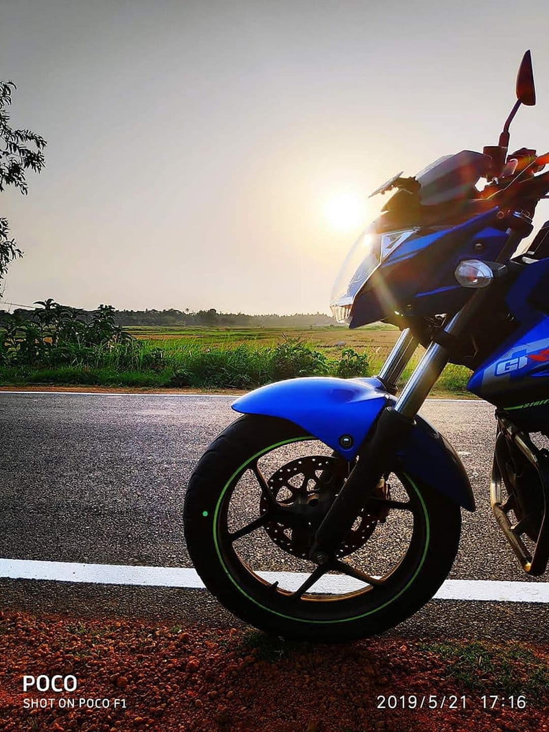 Blue motorcycle parked on a paved road during a golden sunset with lush green fields stretching to the horizon.