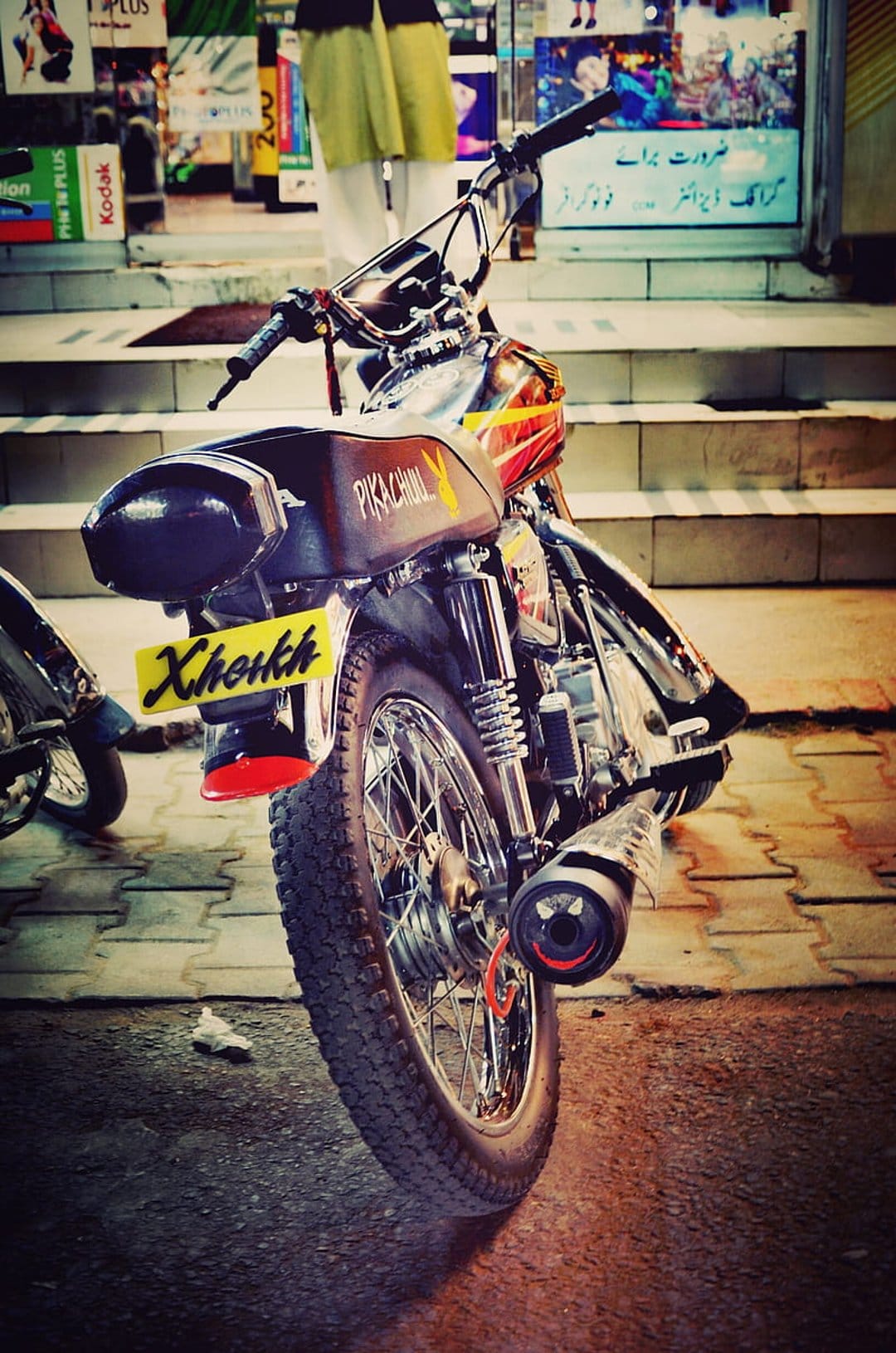 Motorcycle rear featuring a yellow Xkerkh license plate and Pikachu tank logo against a storefront background.