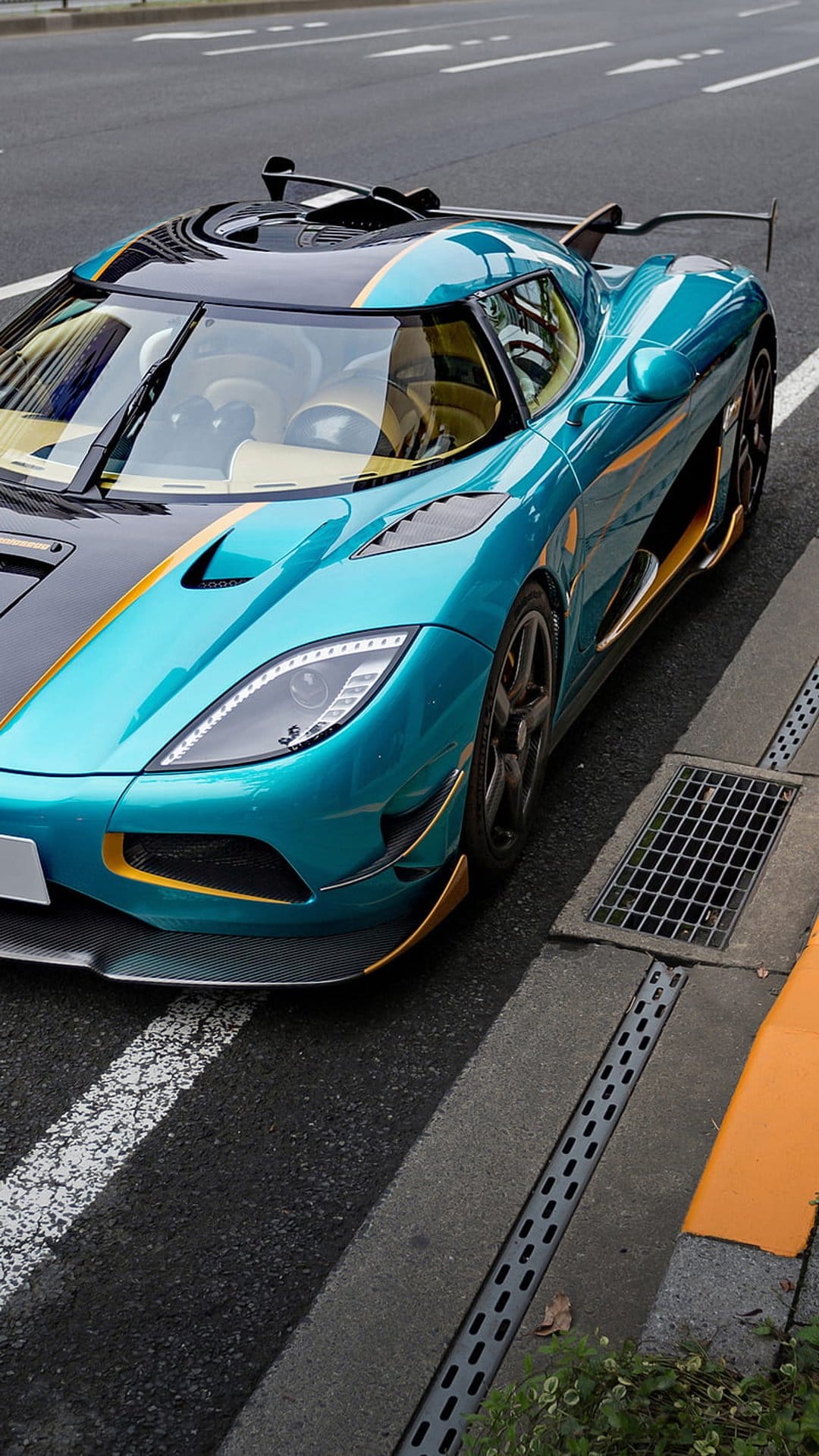 Teal Koenigsegg Agera RS with gold accents parked on an asphalt road next to a yellow curb and metal grate.