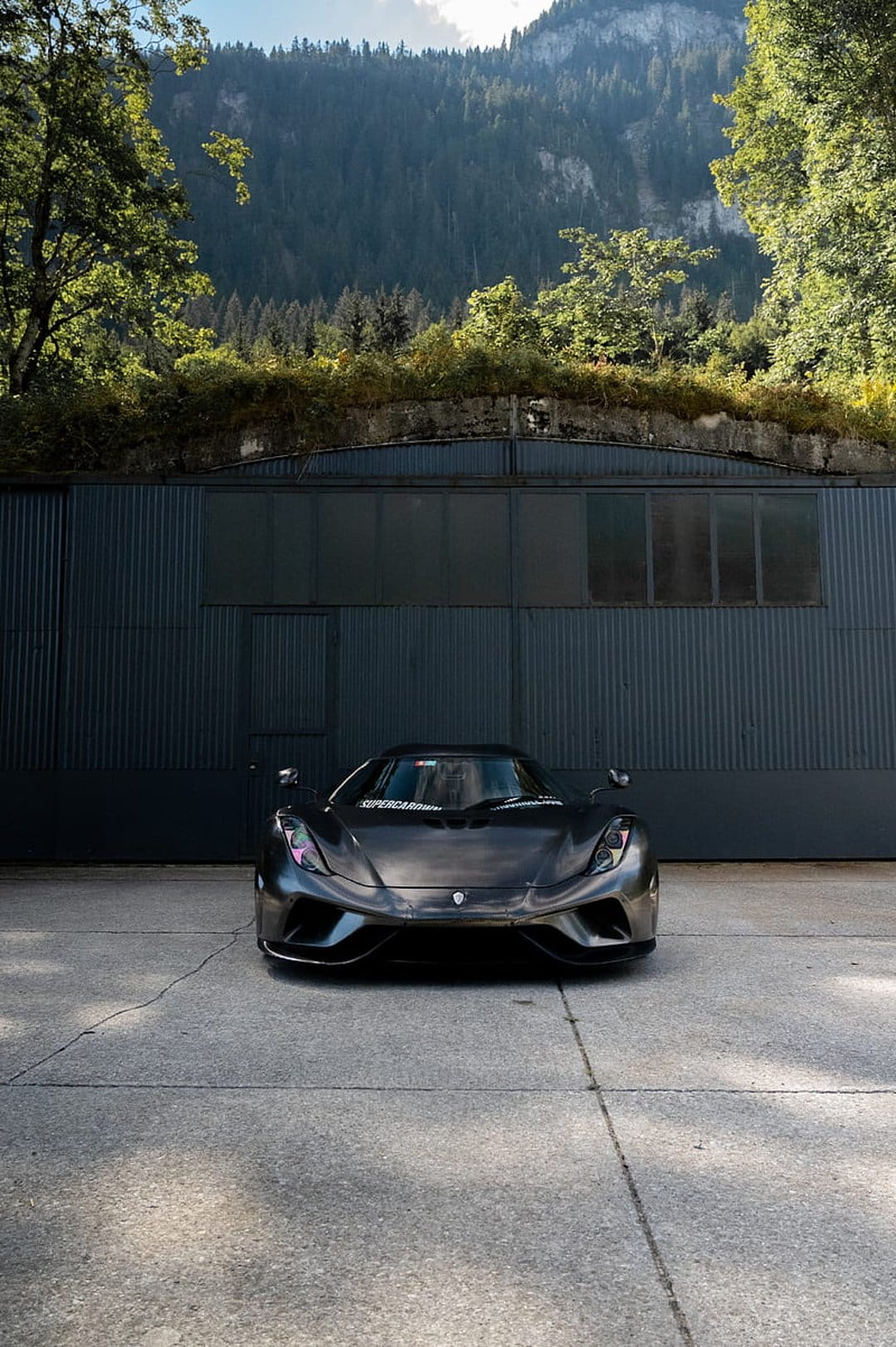 Koenigsegg Jesko hypercar on concrete before an industrial building with forested mountains in the background.