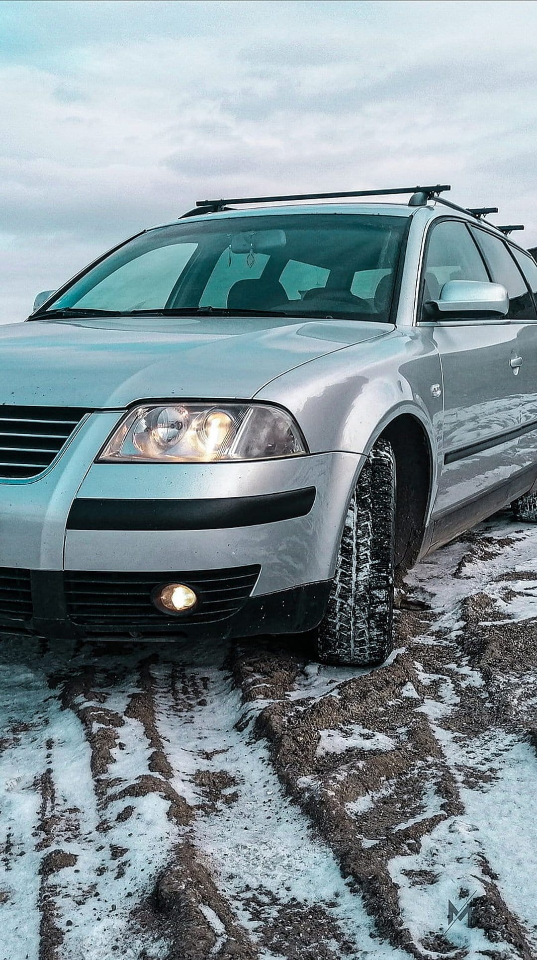 Silver Volkswagen Passat station wagon with glowing headlights parked on a muddy, snow-covered dirt road at dusk.