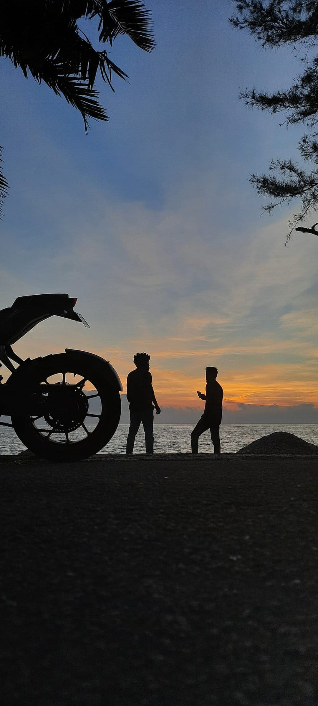 Two friends and a motorcycle stand silhouetted against a vibrant orange sunset on a tropical sandy coastline.