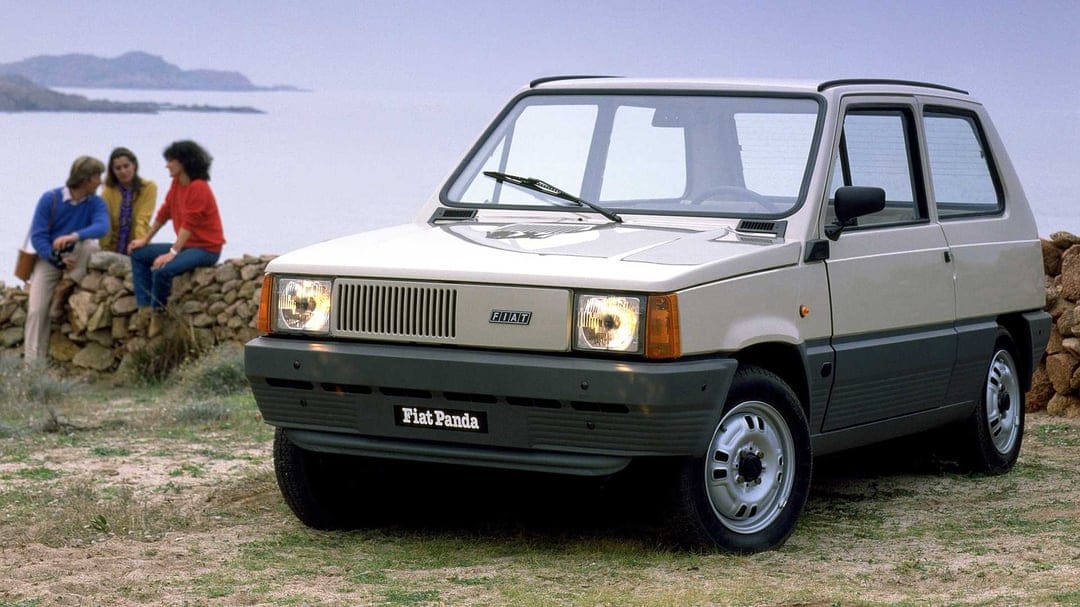 Classic beige Fiat Panda parked on a dirt road near the ocean with people sitting on a stone wall in the distance.