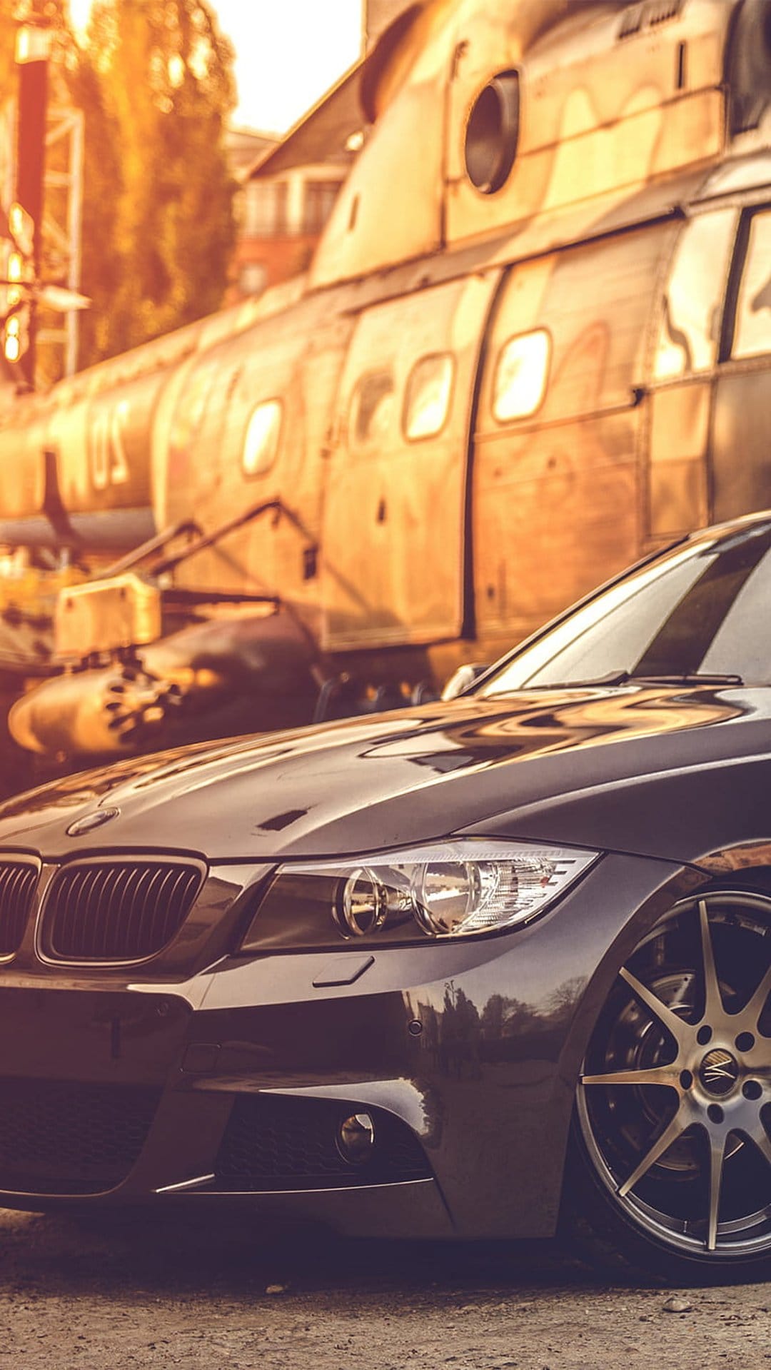 Black BMW car parked beside a large weathered vintage helicopter during a dramatic golden hour sunset outdoors.