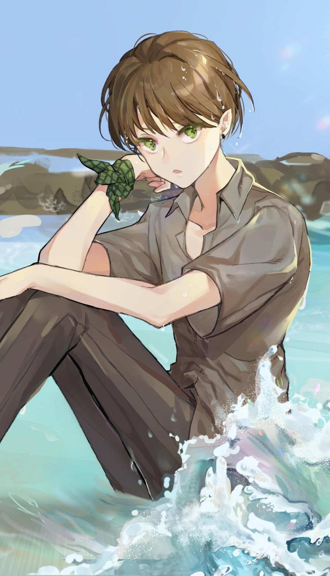 Brown-haired young man with green eyes sits on a beach while ocean waves crash around his gray shirt and dark pants.