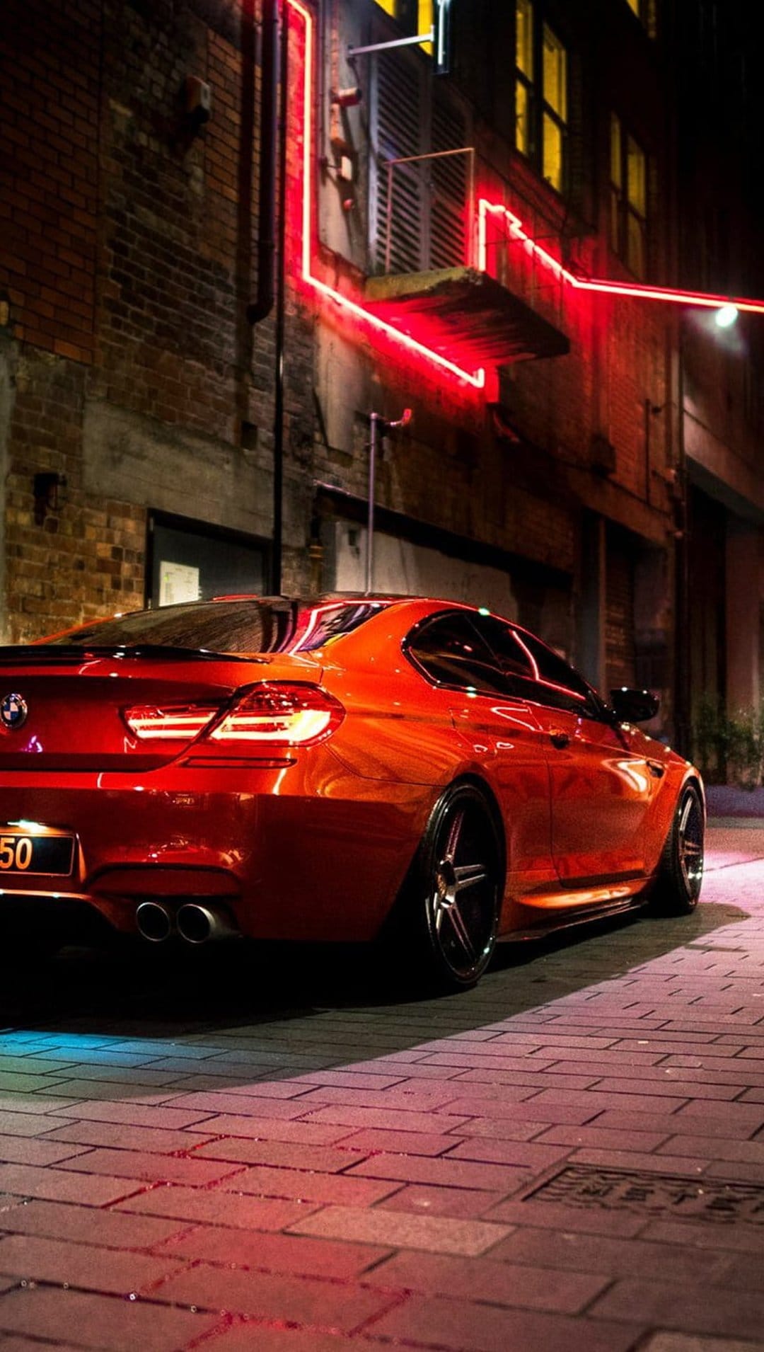 Crimson BMW coupe on wet bricks under red neon lighting with dramatic reflections on the car and pavement.