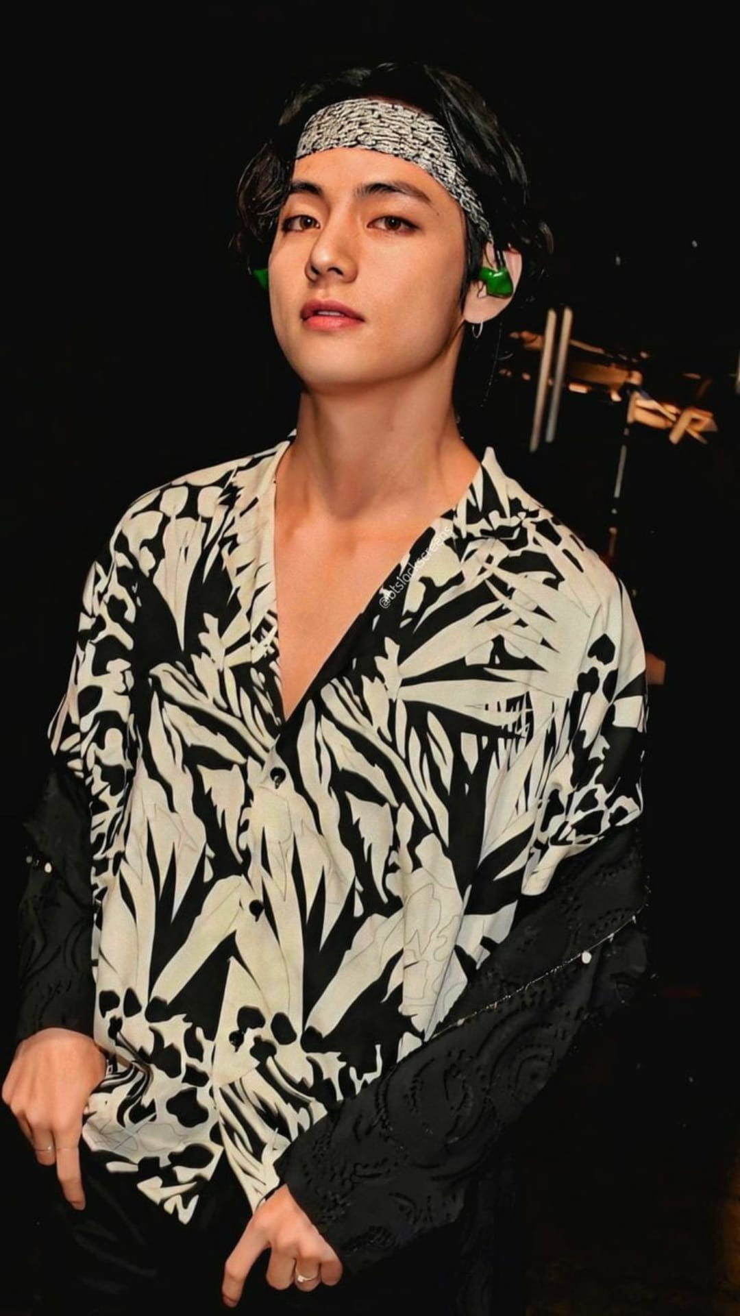Kim Taehyung of BTS wearing a black and white leaf-print shirt, a dark bandana, and small green earbuds.