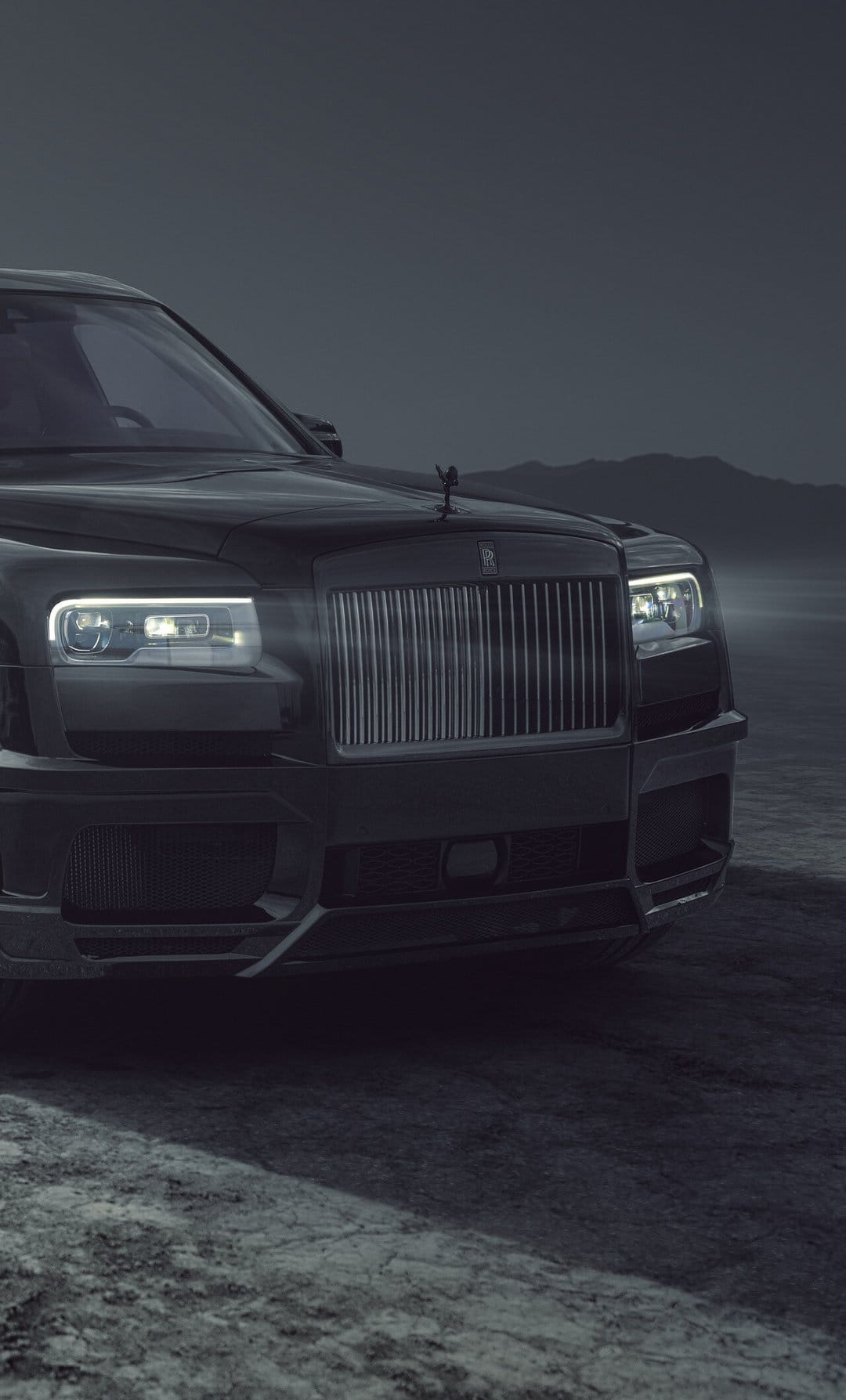 Close-up of a black luxury SUV front grille and glowing headlights in a misty, dark mountain landscape.