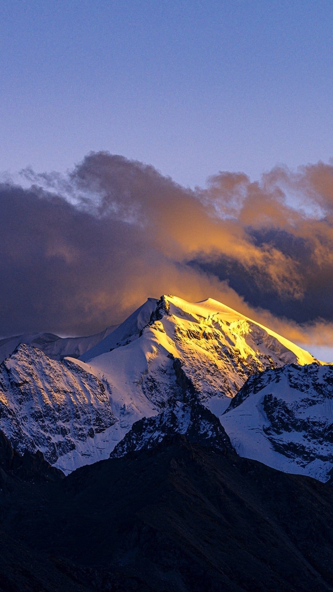 Snow-capped mountain summit glowing gold under a purple twilight sky with dramatic shadowed clouds at sunset.