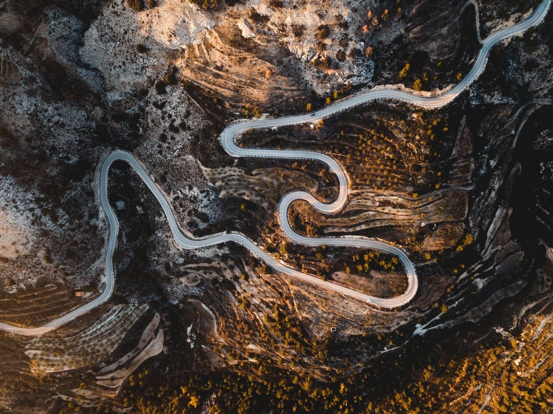 Serpentine asphalt road weaves through rugged mountains as golden sunset light creates long shadows on the rocks.