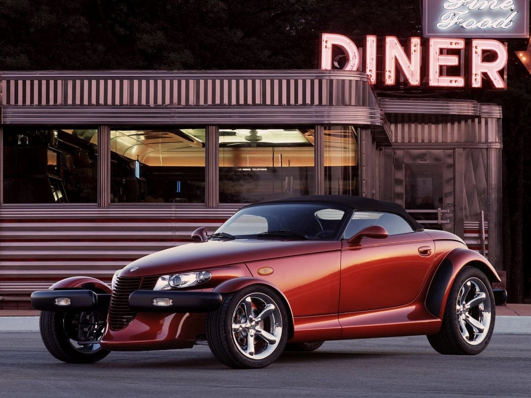 Copper Plymouth Prowler parked in front of a retro diner with a red neon sign at night with warm reflections.