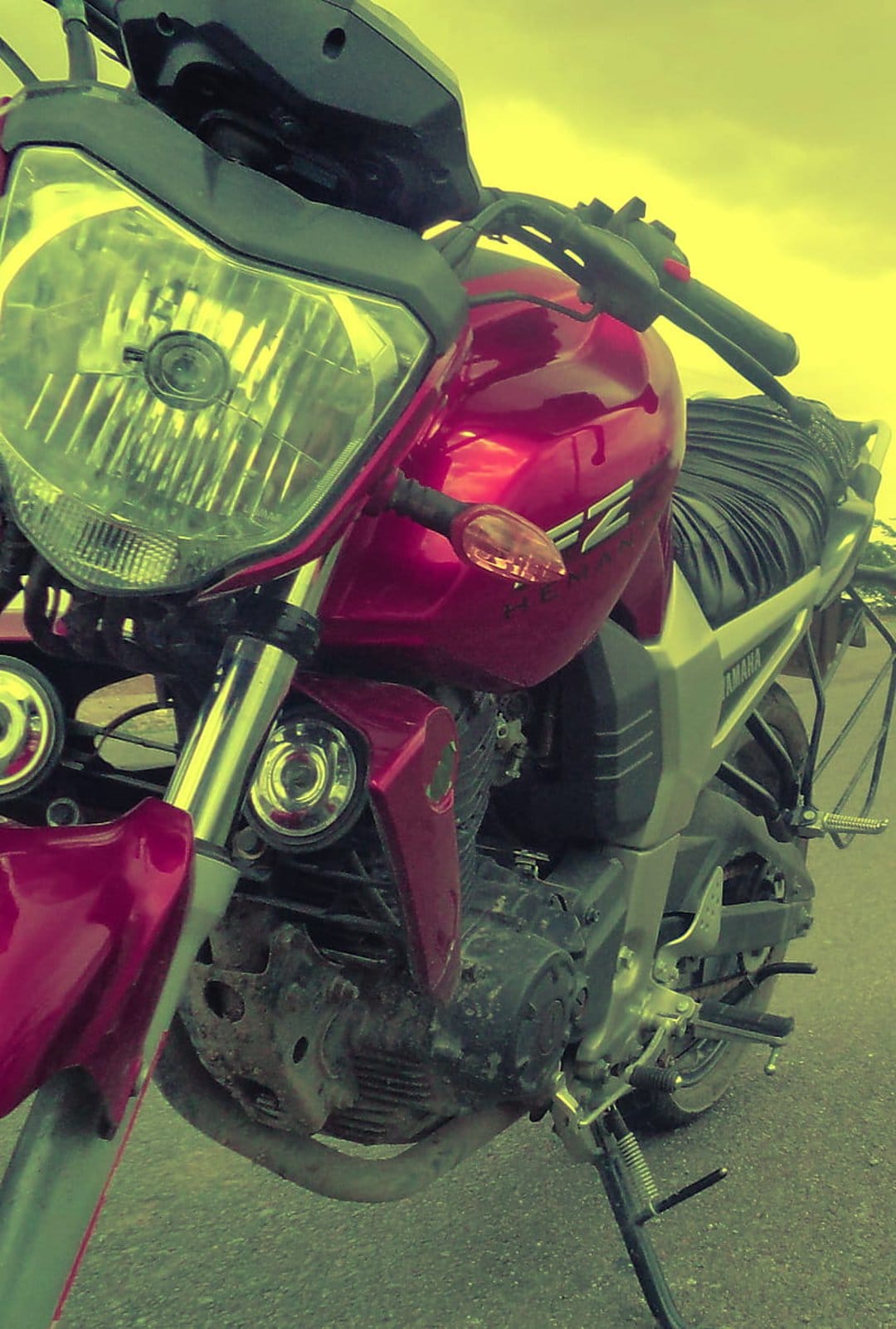 Red Yamaha motorcycle shows the front headlight, handlebars, and textured seat against a muted yellow sky.