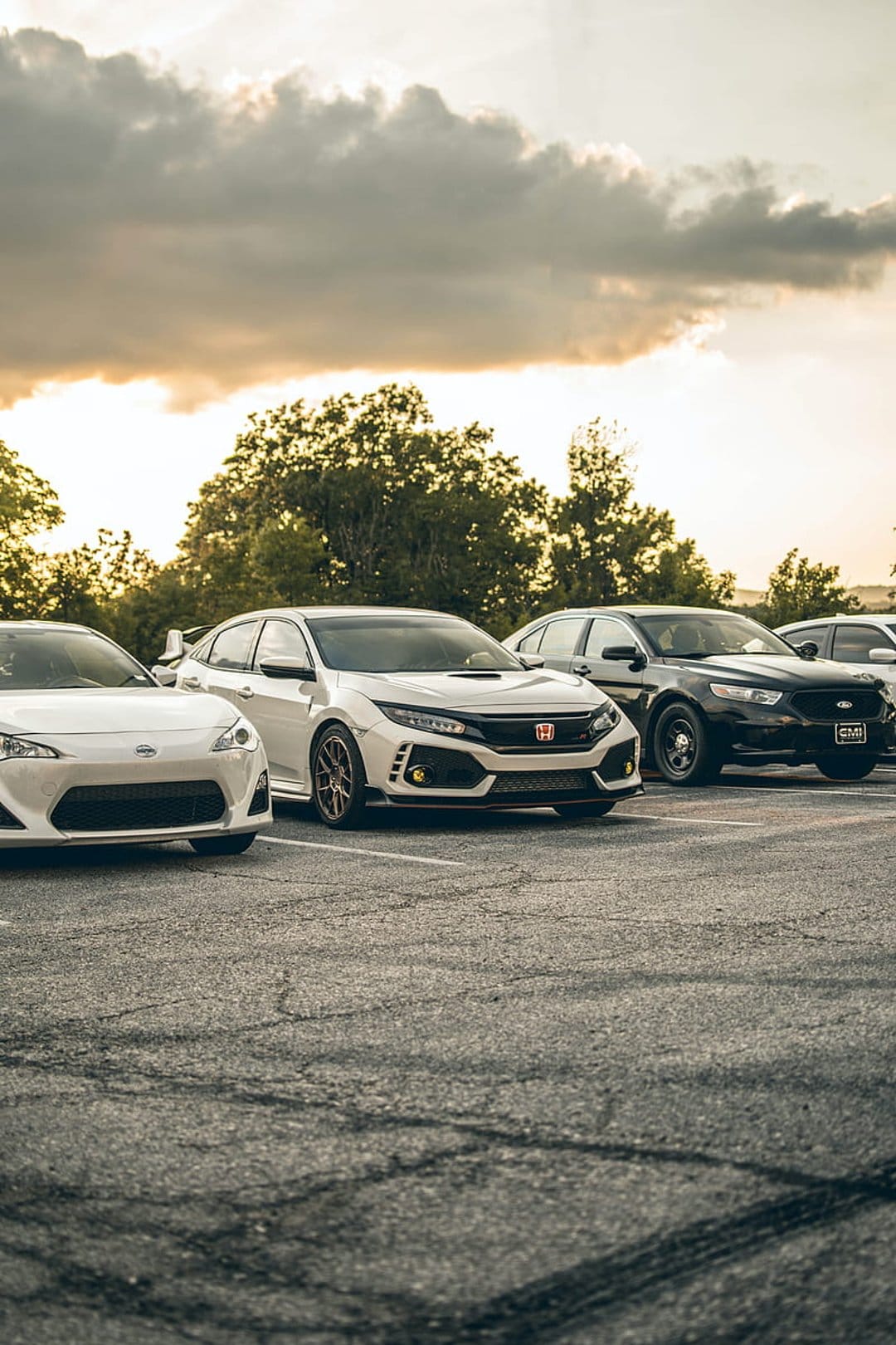 Sleek white and black cars parked on asphalt beneath a dramatic sunset sky filled with dark, heavy storm clouds.