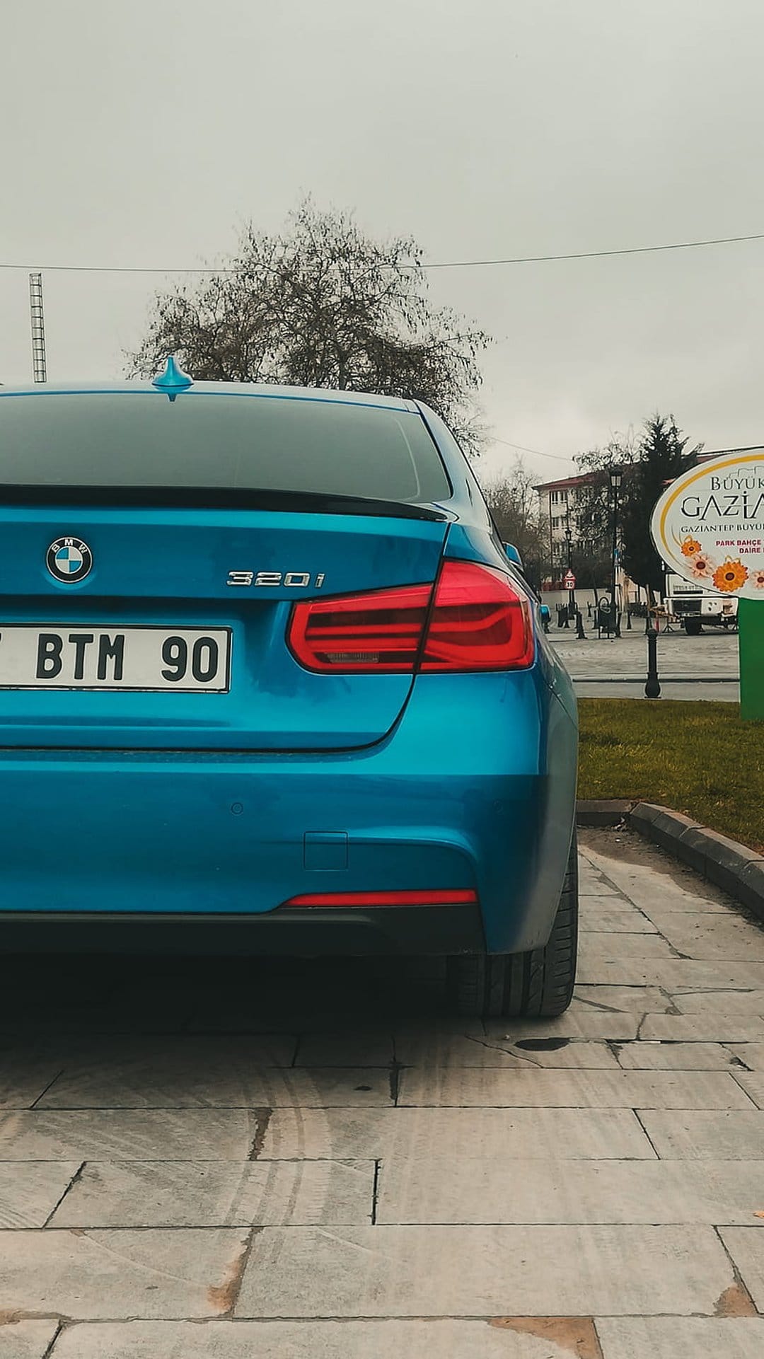 Blue BMW 320i sedan rear view parked on stone pavers with tire marks under a cloudy sky and bare tree branches.