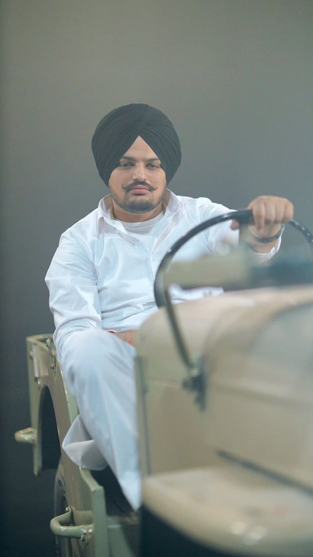 Man in a black turban and white kurta holds the steering wheel of a vintage jeep against a gray wall.