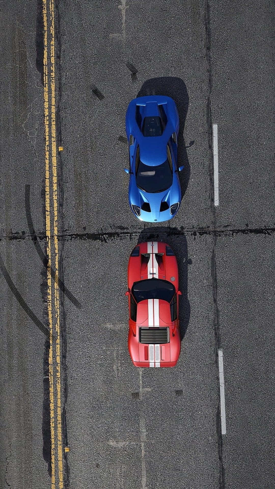 Red and blue sports cars race side-by-side on a textured asphalt road from a direct overhead aerial viewpoint.