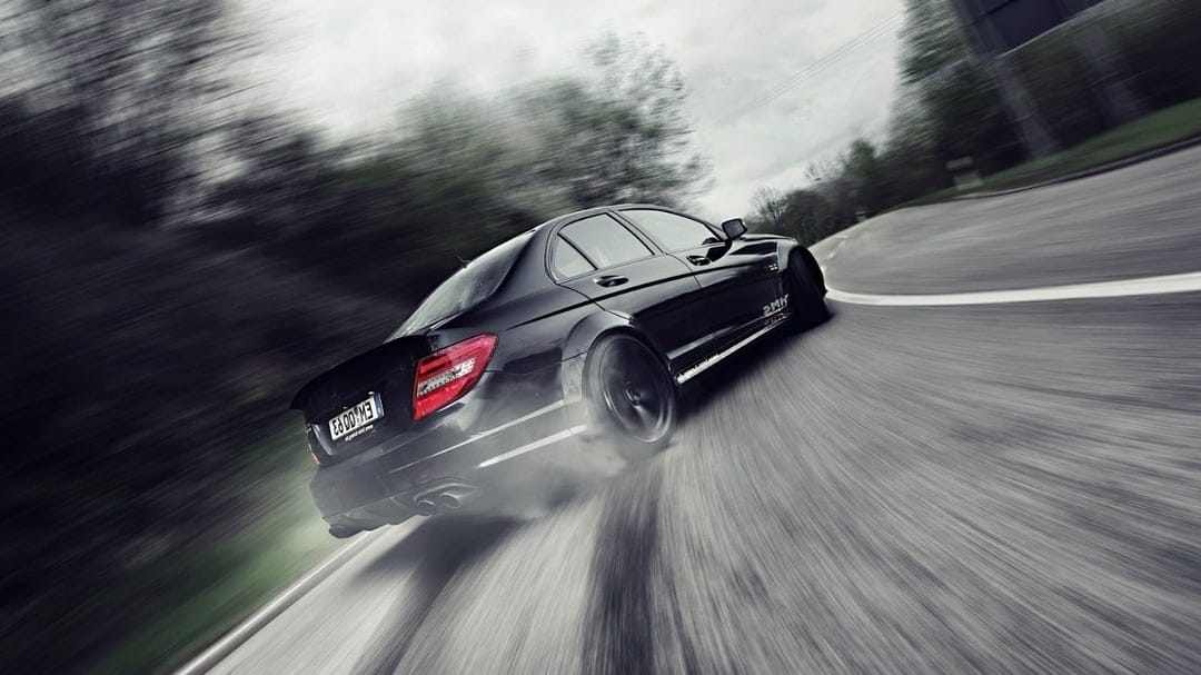 Black Mercedes-Benz C63 AMG drifting on a curved road with thick tire smoke and motion-blurred background.