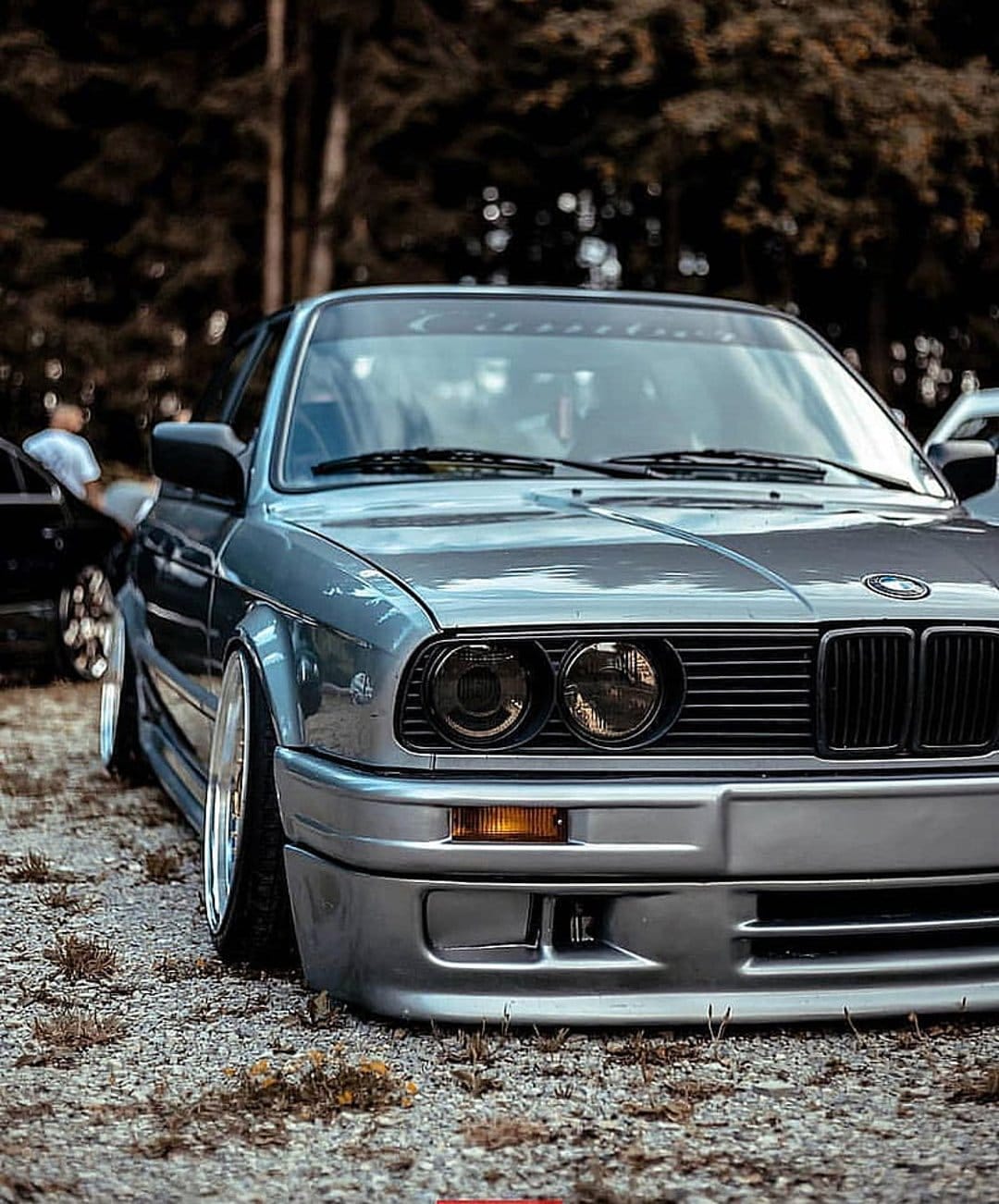 Silver BMW E30 coupe with a lowered stance parked on a gravel road with blurred green trees in the background.