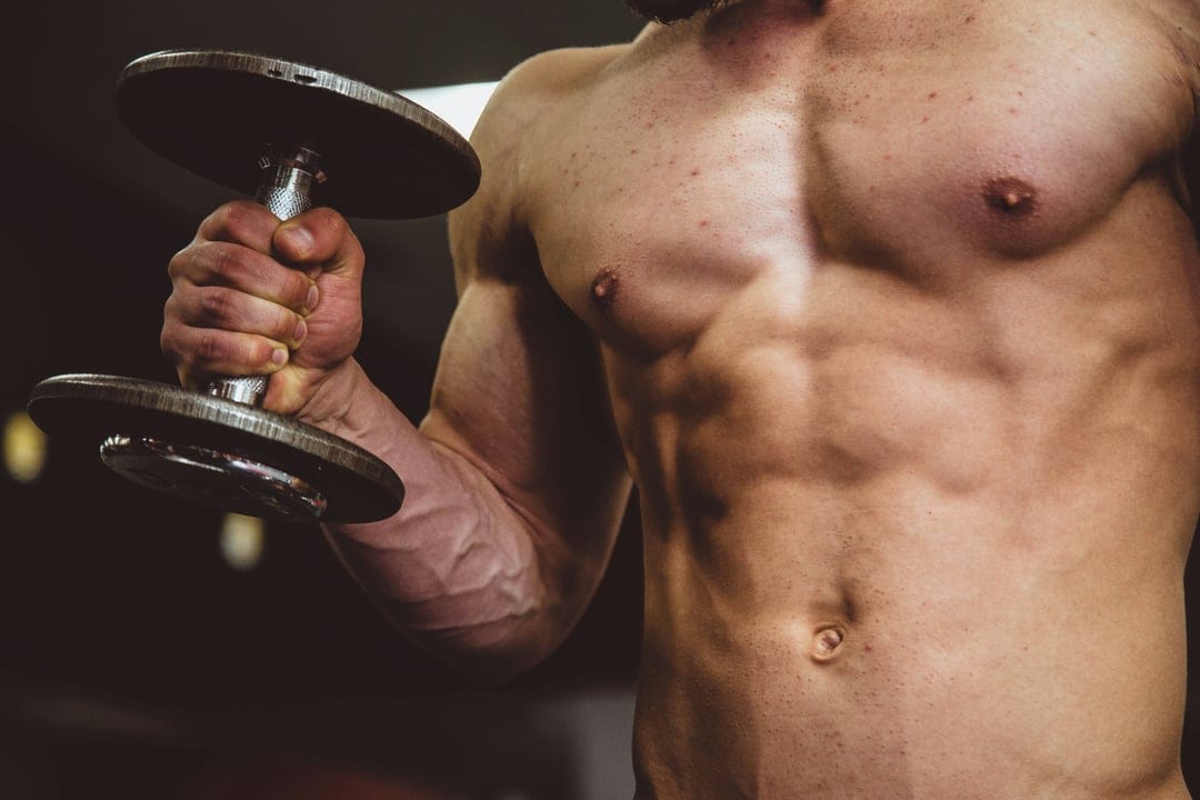 Muscular man with defined biceps and vascular arms lifts a dumbbell, highlighting his physique in a dark studio.