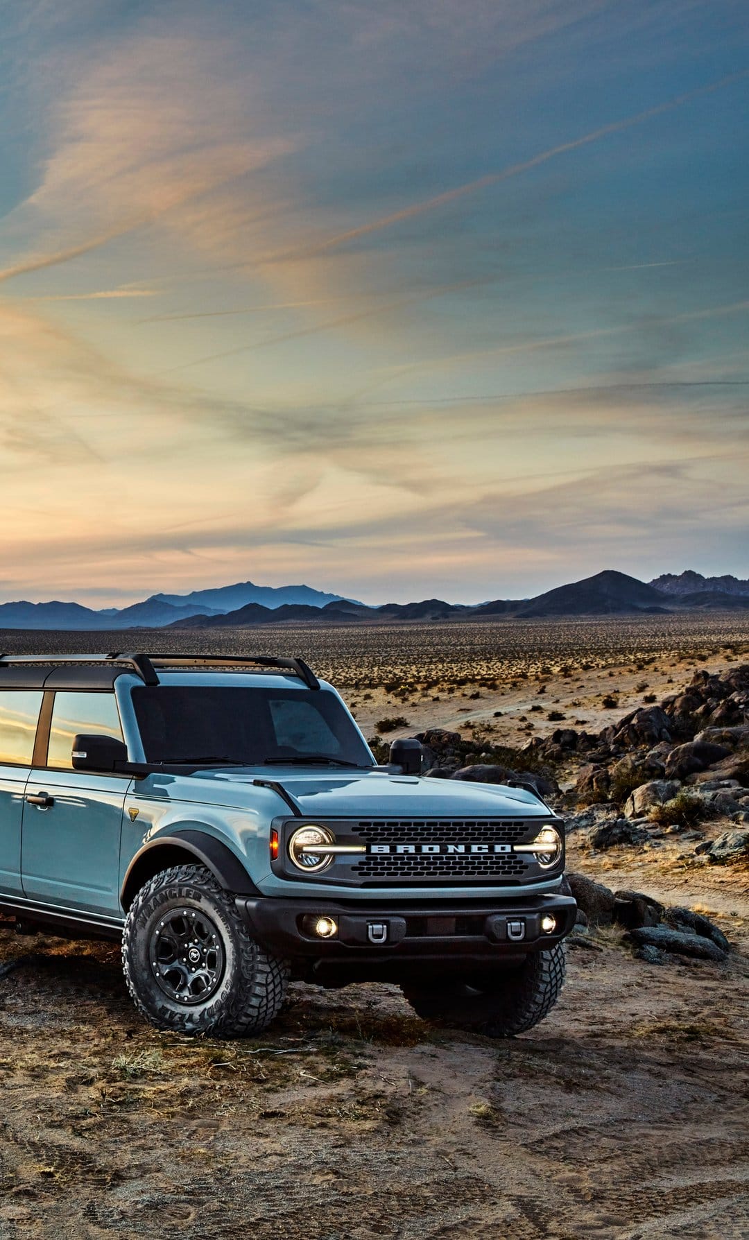 Blue Ford Bronco parked on rocky desert ground under a soft orange and blue sunset sky with distant mountains.