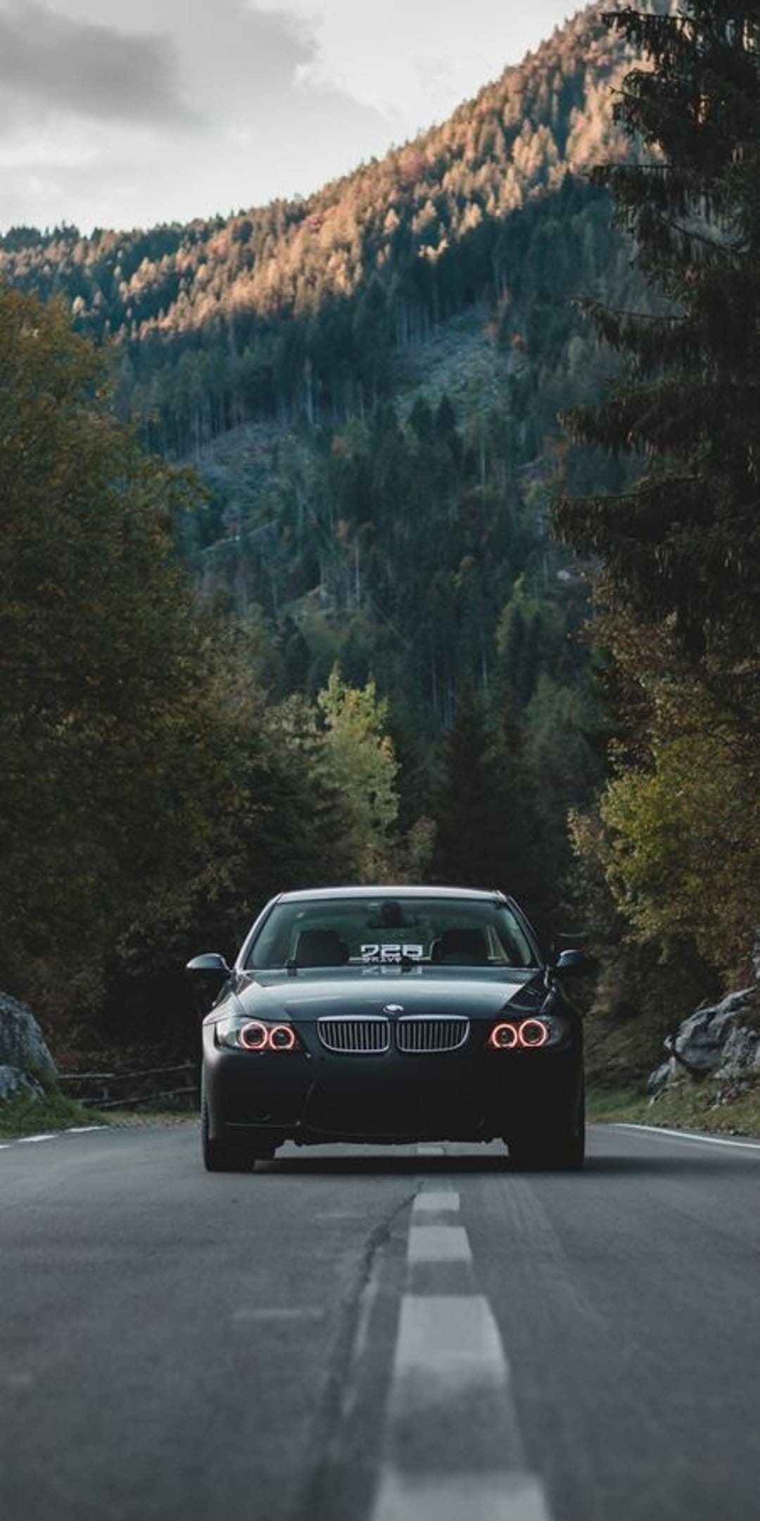 Dark BMW M3 with glowing headlights drives on a winding road through a dense forest with autumn colors and mountains.
