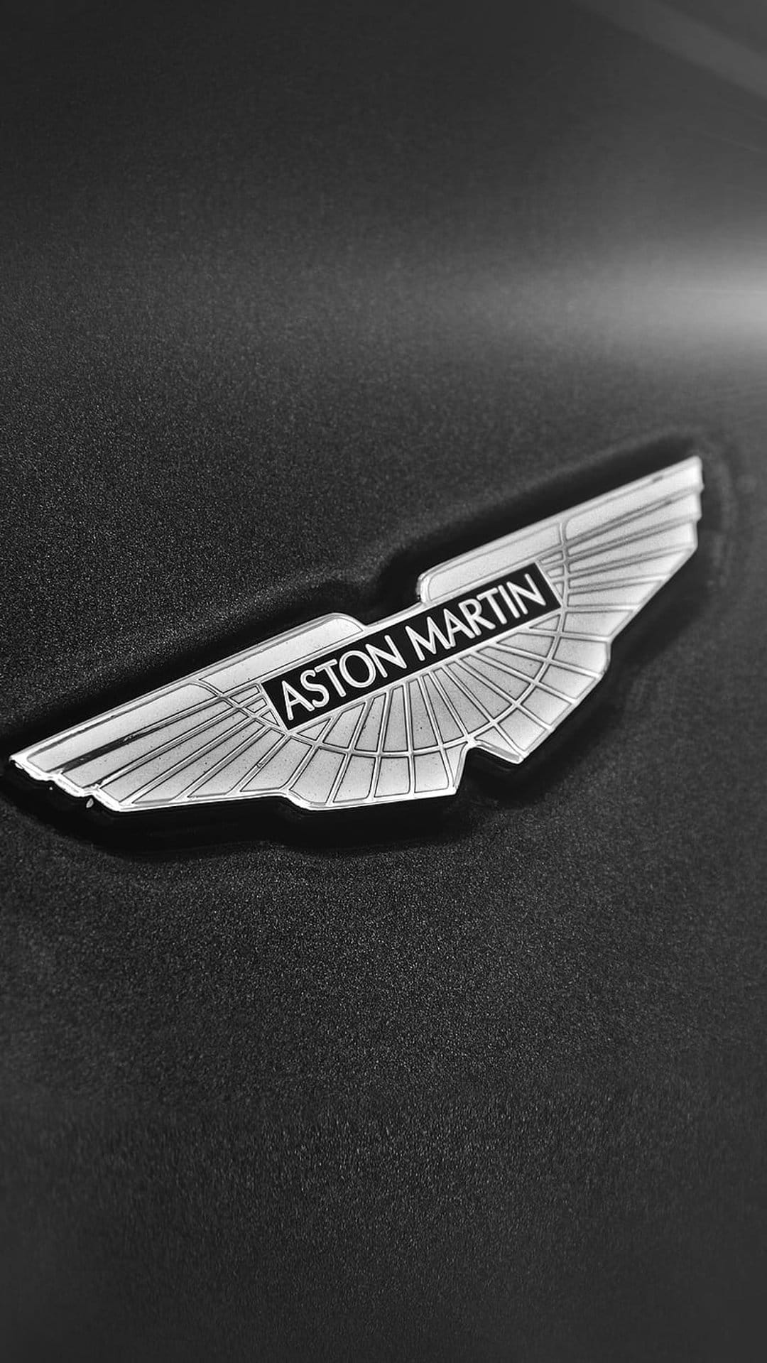 Aston Martin wings logo in silver metal centered on a dark grainy surface with high contrast monochrome lighting.
