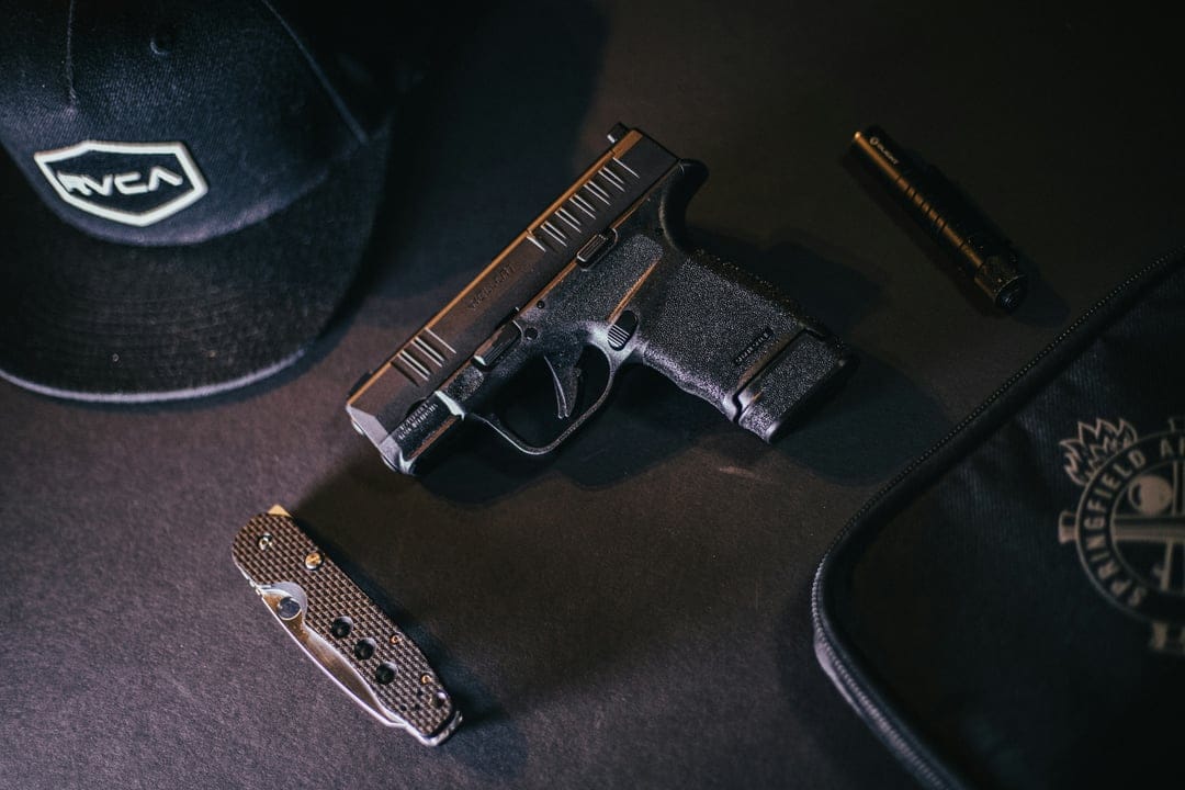 Black semi-automatic pistol, folding pocket knife, and tactical cap arranged on a dark equipment case background.