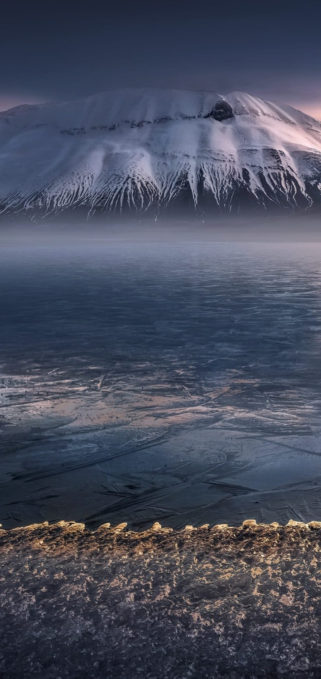 Jagged snow-covered mountains rise behind a frozen glacial foreground under a deep blue and purple twilight sky.