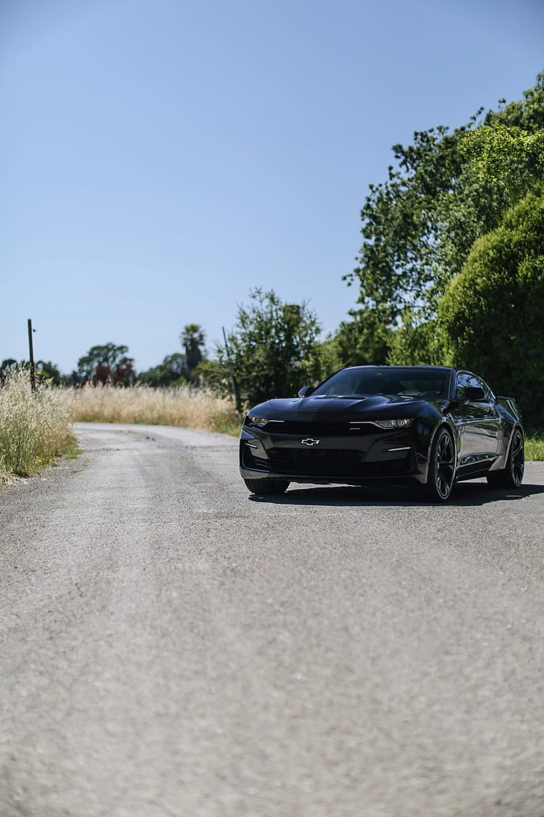 Black Chevrolet Camaro drives on a dusty road surrounded by green trees and tall grass under a clear blue sky.