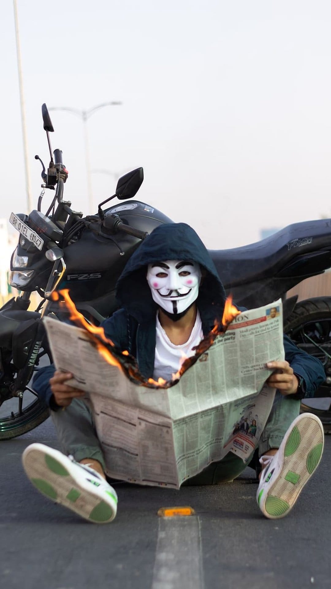 Masked figure in a Guy Fawkes mask burns a newspaper while sitting on asphalt next to a motorcycle at night.