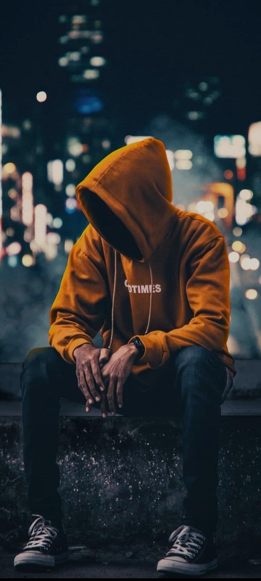 Man in an orange hoodie sits on a ledge overlooking glowing city lights during a dark urban night scene.