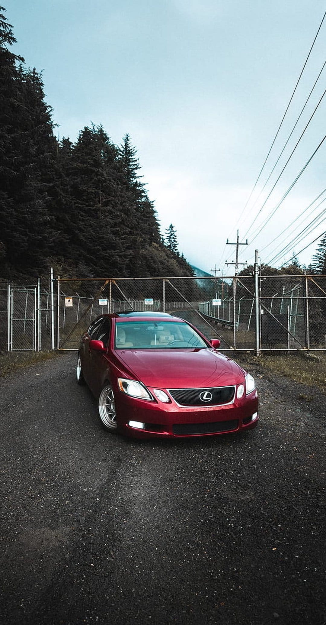 Red Lexus GS luxury sedan parked on a gravel road before a gated pine forest under a moody overcast sky.