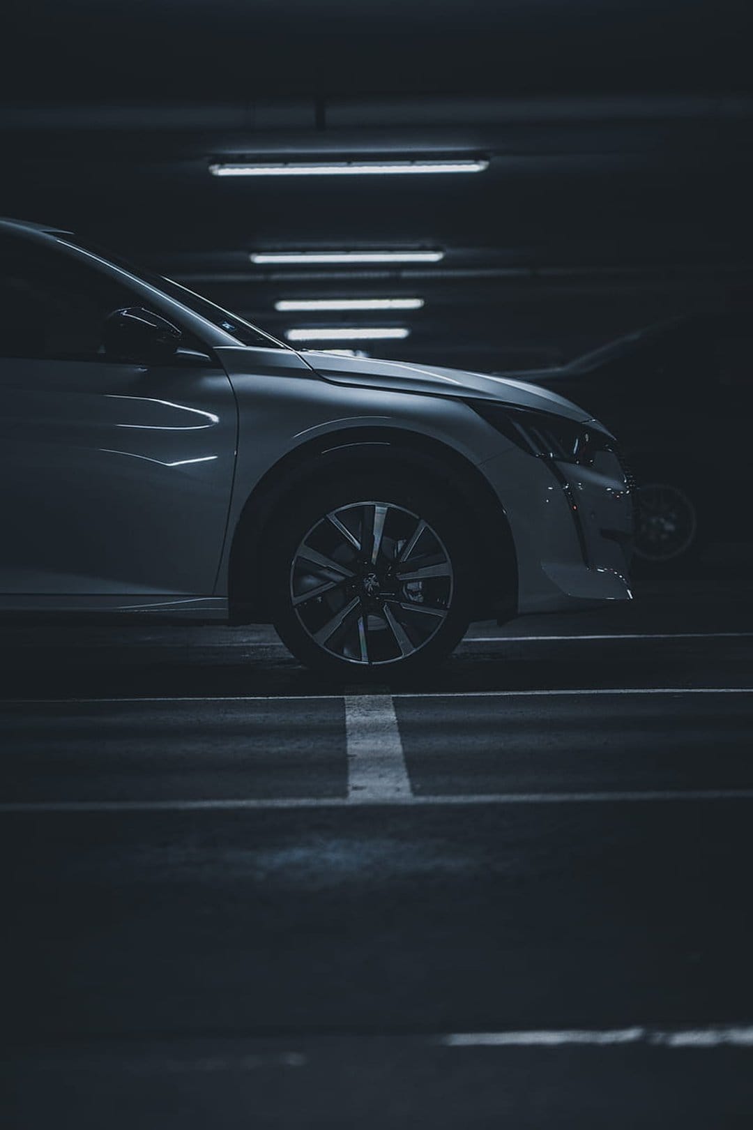 Modern car front wheel and fender illuminated by overhead fluorescent lights in a dark, shadowy parking garage.