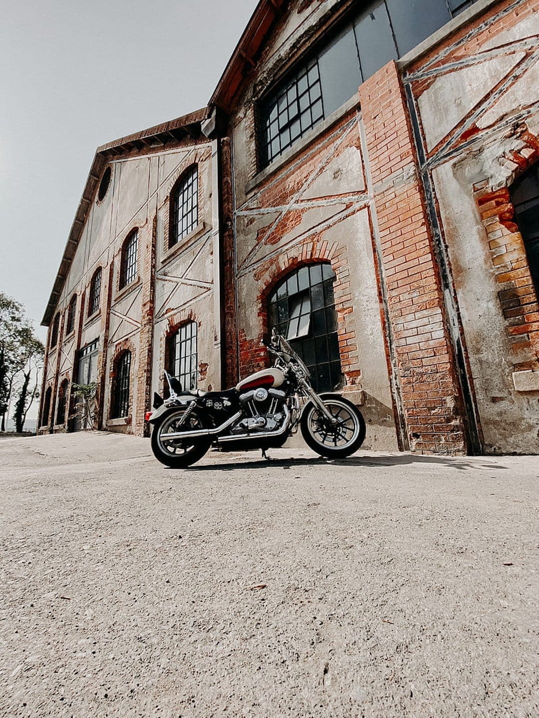 Red and white motorcycle on gravel in front of an industrial brick building with large multi-paned windows.