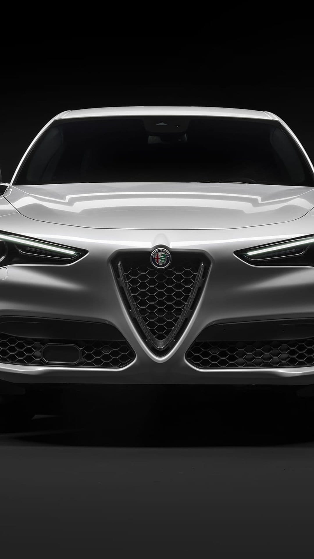 White Alfa Romeo Stelvio SUV front view with illuminated LED headlights and shield grille on a black background.