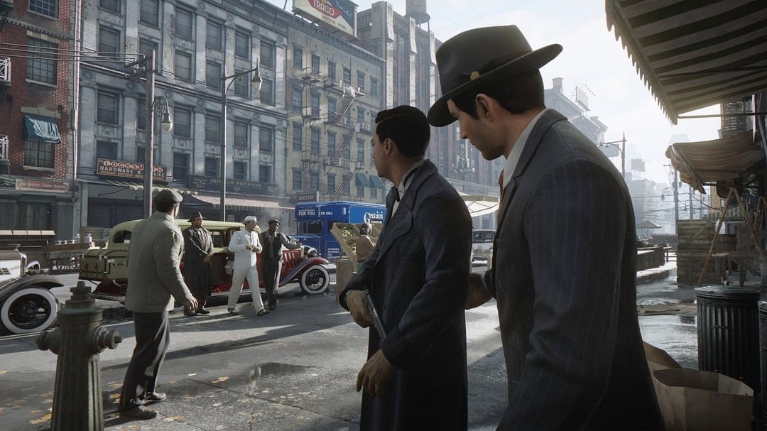 Two men in suits stand on a 1930s city street with vintage cars and old brick buildings in the background.