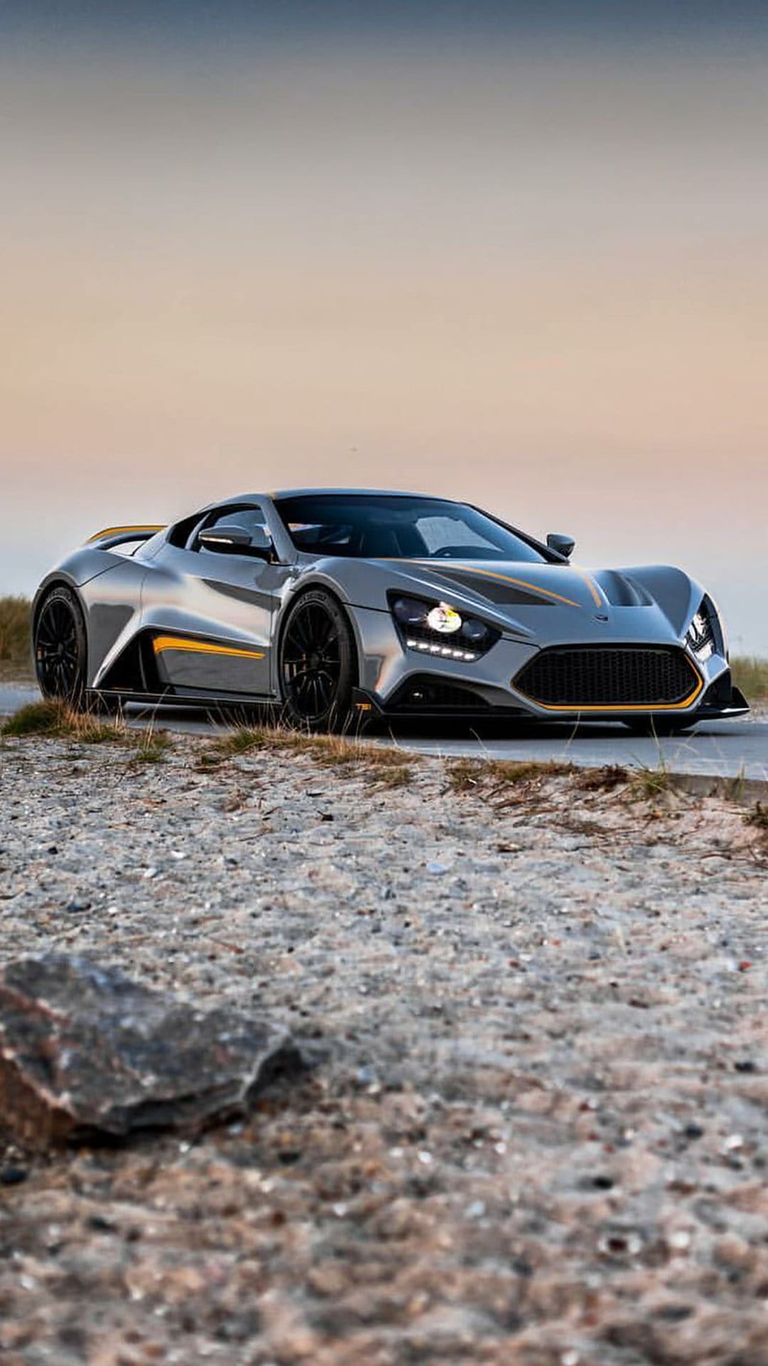 Silver Zenvo TSR-S hypercar with yellow accents parked on a gravel coastal road during a soft sunset.