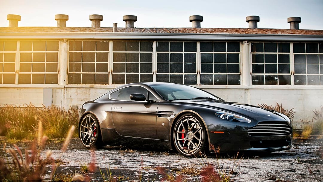 Dark gray Aston Martin supercar parked in front of an industrial building during a golden hour sunset.