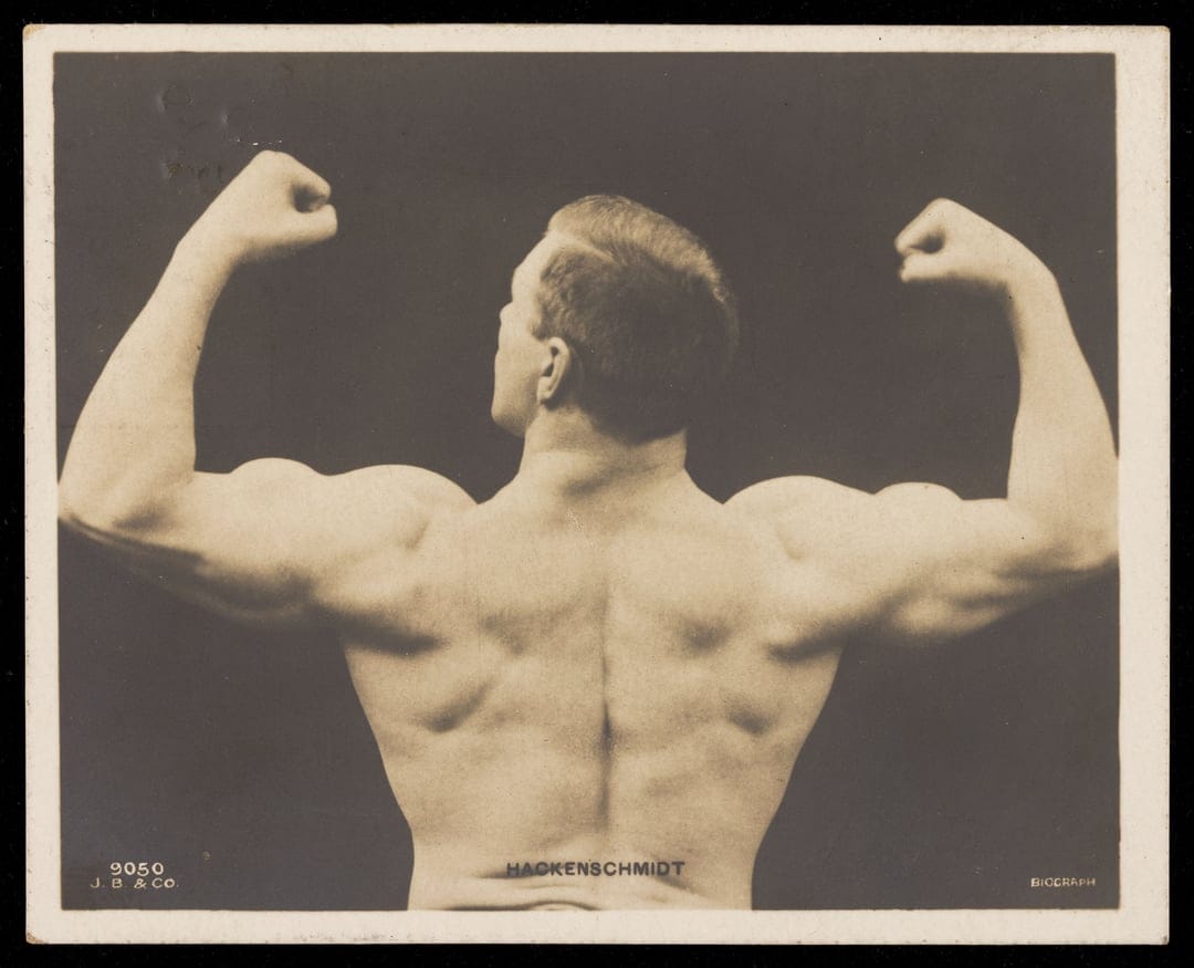 George Hackenschmidt flexes his muscular back and biceps in a sepia-toned vintage portrait against a dark background.