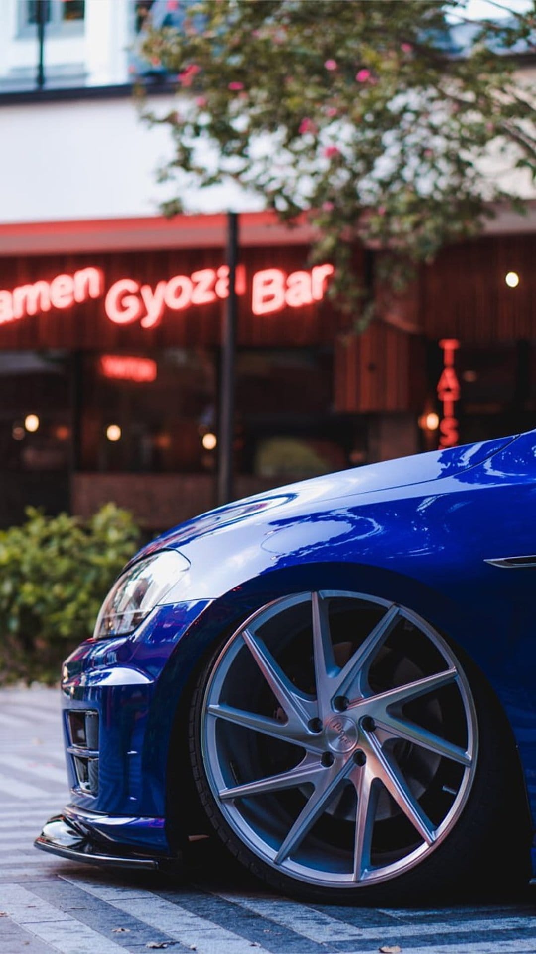 Blue sports car with silver rims parked on cobblestones next to a red neon Ramen Gyoza Bar sign at night.