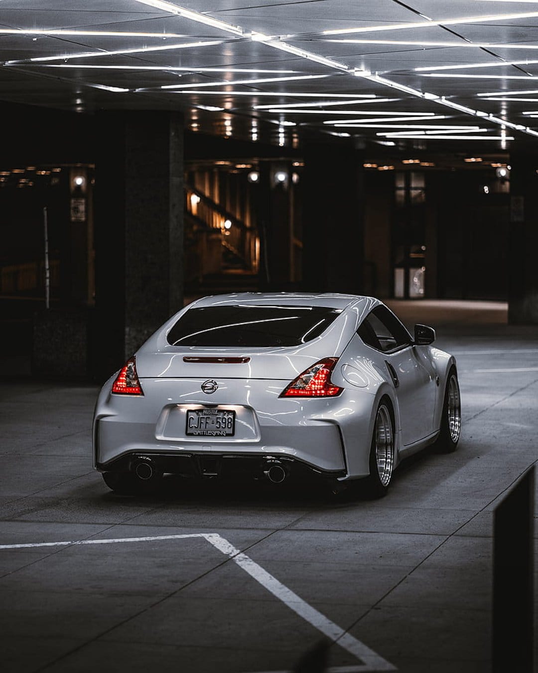 Nissan 370Z rear view with lit red taillights parked under linear ceiling lamps in a dark concrete parking garage.