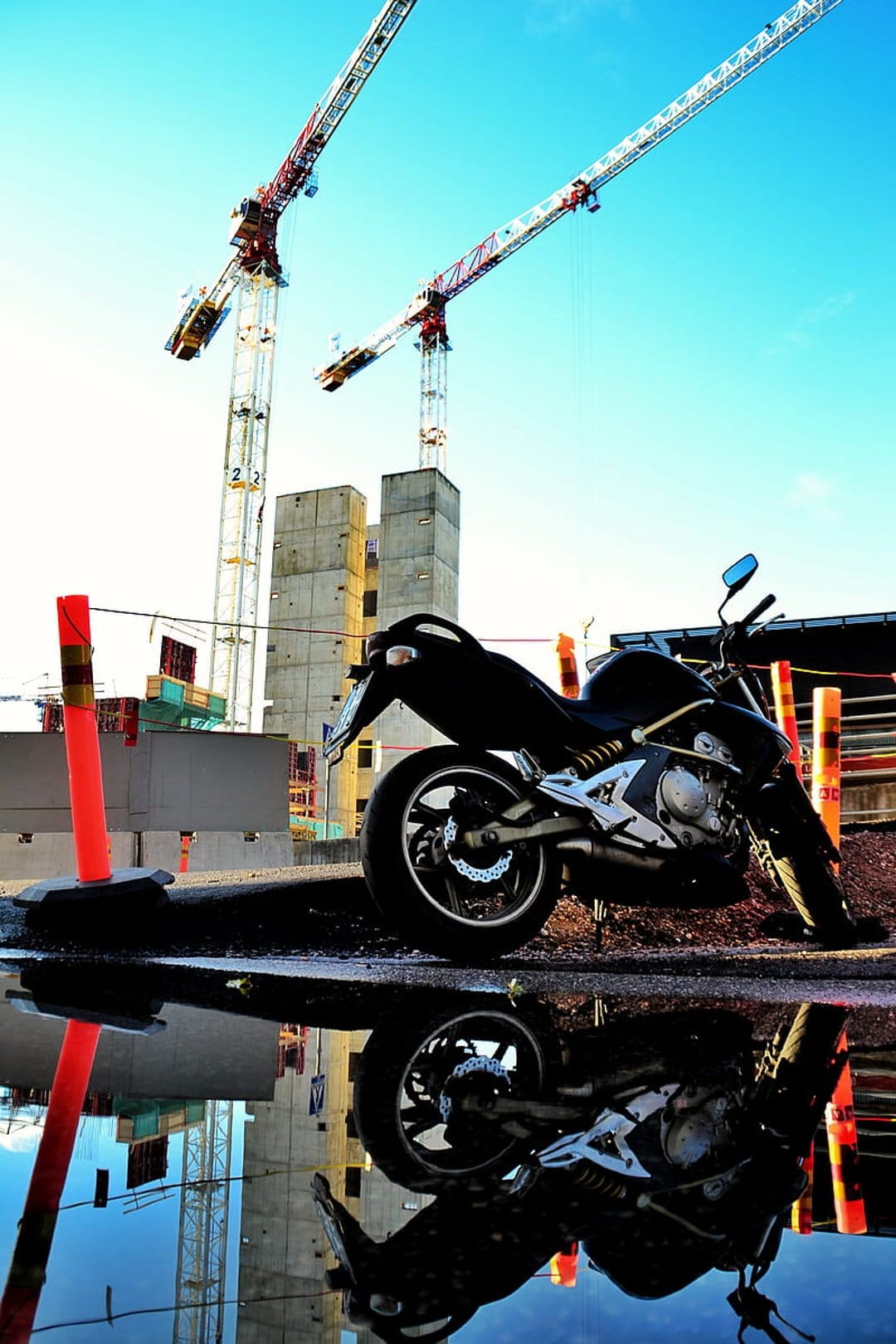 Black motorcycle reflecting in a street puddle with industrial cranes and a clear blue sky in the background.