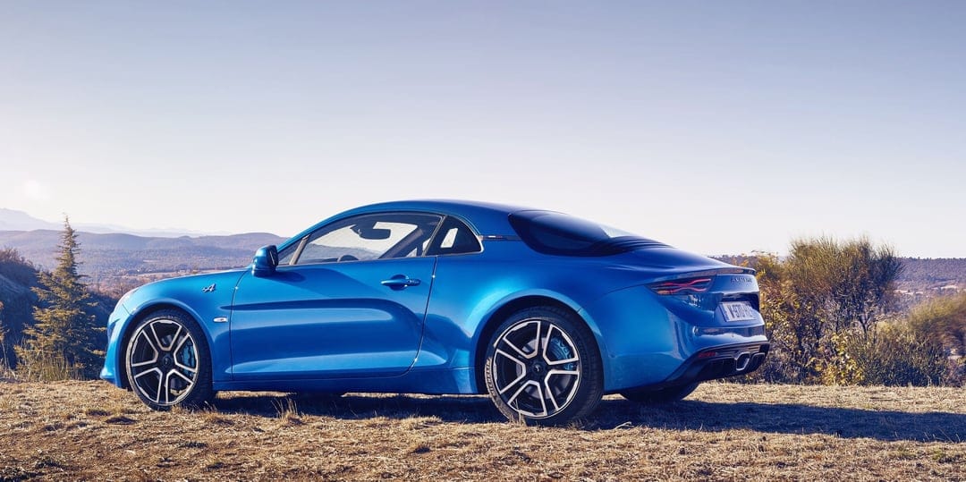 Blue Alpine A110 sports car side profile on a grassy hill with mountains and a clear sky in the background.