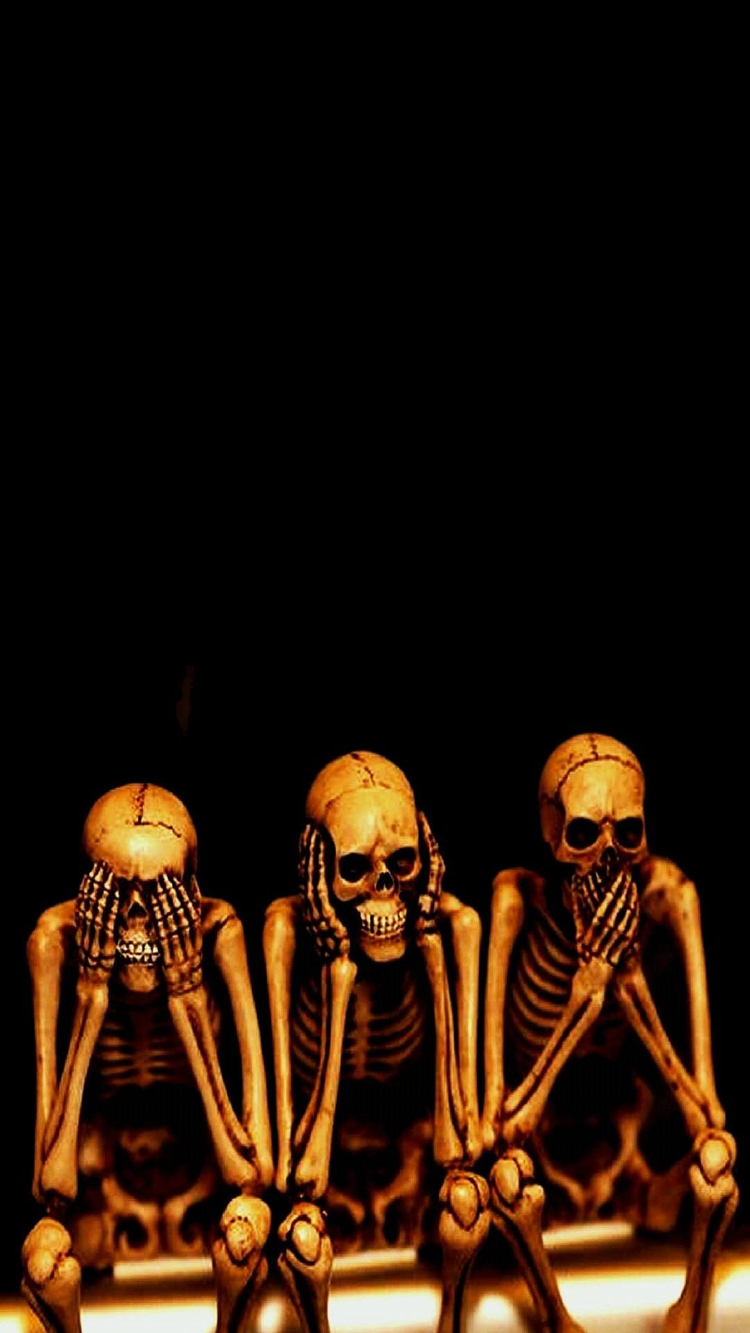 Three skeletons sit in a row covering their eyes, ears, and mouth with bony hands under warm spotlighting.
