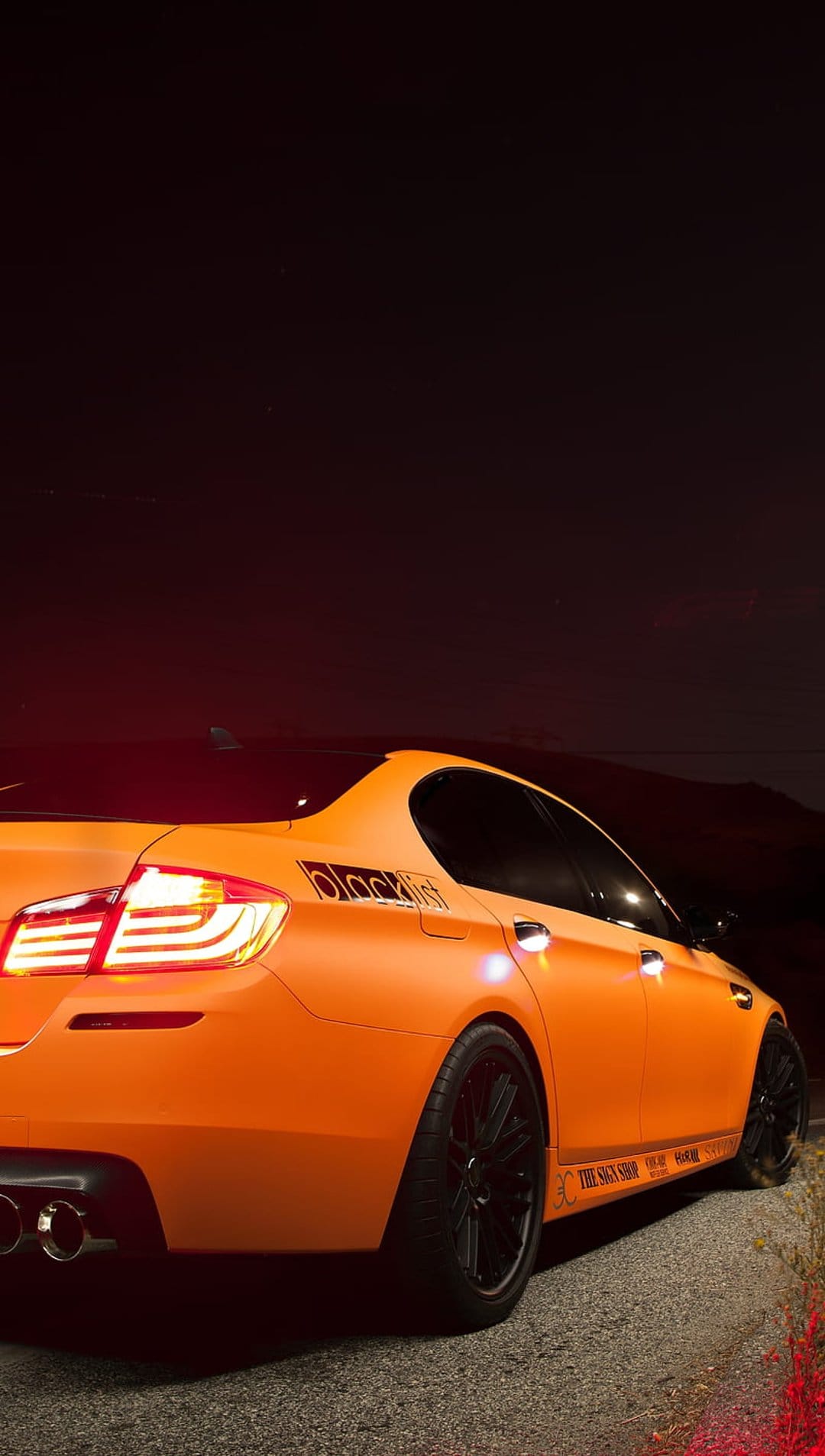 BMW M5 sedan with matte orange finish and black rims parked on a dark street with glowing red taillights.